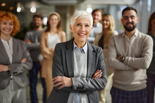 Happy mature business leader standing with crossed arms in front of her team in the office and looking at camera.