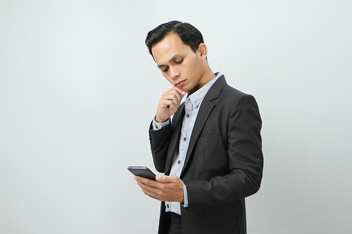 pensive asian indonesian business man in suit holding smart phone on isolated background