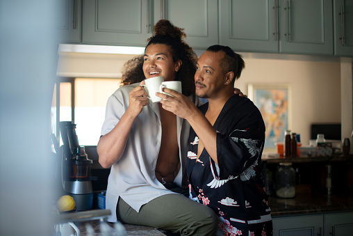Coffee, cheers and gay couple in kitchen together at modern home for bonding and talking in morning. Love, conversation and young lgbtq men in relationship drinking cappuccino or latte at apartment.