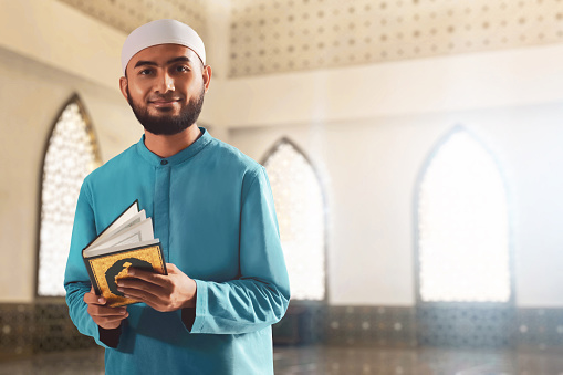 Portrait of handsome young asian muslim man with beard posing,  holding holy book quran and smiling in the mosque window arch