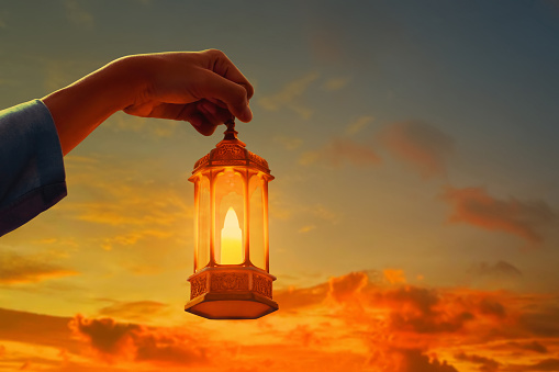 Hands of young asian muslim man holding shiny arabic lantern at beautiful sunset sky with cloud