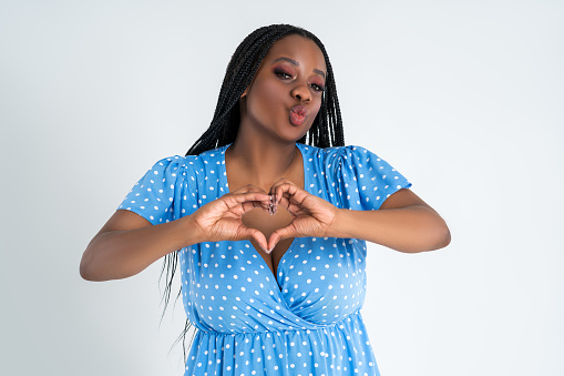 Happy plus size female model posing in blue dress on gray studio background and showing heart sign, young African woman with curvy figure and pigtailed hairstyle, afro braids