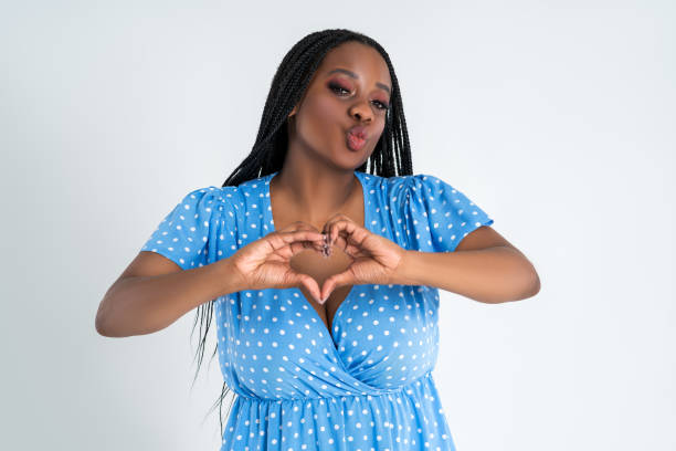 Happy plus size female model posing in blue dress on gray background and showing heart sign, young African woman with curvy figure and pigtailed hairstyle, afro braids Happy plus size female model posing in blue dress on gray studio background and showing heart sign, young African woman with curvy figure and pigtailed hairstyle, afro braids big fat lips stock pictures, royalty-free photos & images