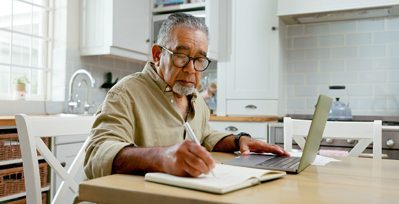 Asian man, mature and kitchen with laptop, notebook. and glasses for notes or taxes. Senior person, computer and technology for communication, work or online banking for retirement and investment