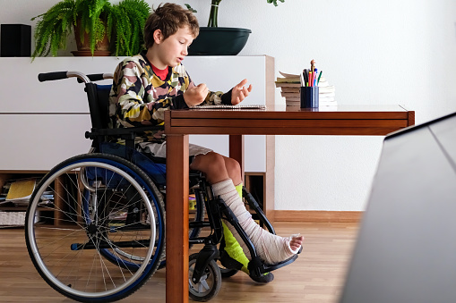 A young boy with a cast on one leg intently focuses on his homework from the comfort of his wheelchair, showcasing determination and adaptability in his home environment.