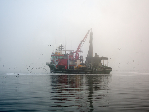 A small fishing vessel backed by a large fog bank sails along the coast