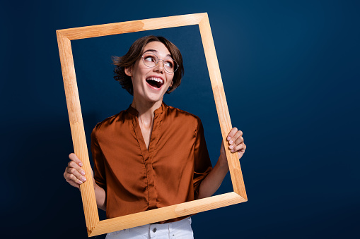Photo portrait of attractive young woman hold wooden frame look empty space wear trendy brown blouse isolated on dark blue color background.
