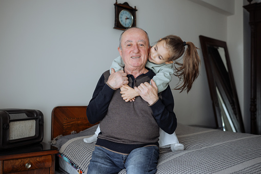 Little cute girl and her grandfather are spending time together at home. Having fun, hugging and smiling while sitting on bed