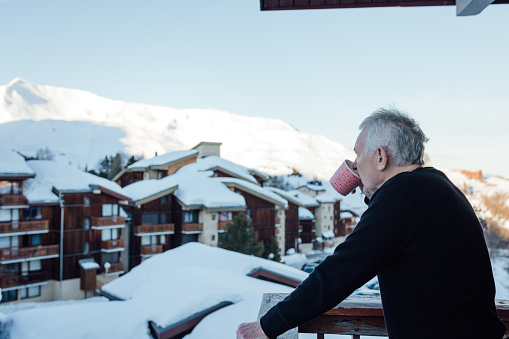 A senior man on vacation at a mountain resort .He is standing on his balcony and enjoying the winter day with a  hot drink. The mountains in the background are covered with snow.