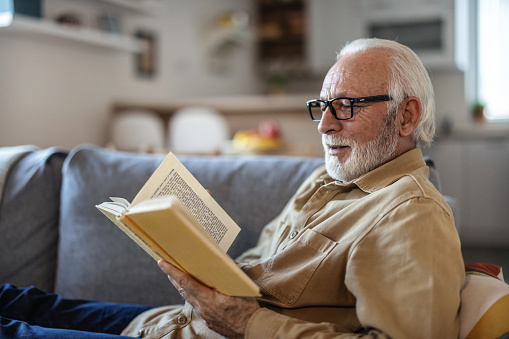 Close up of old happy and relaxed man sitting reading a book at home. Mature male person enjoying free time having fun indoor.