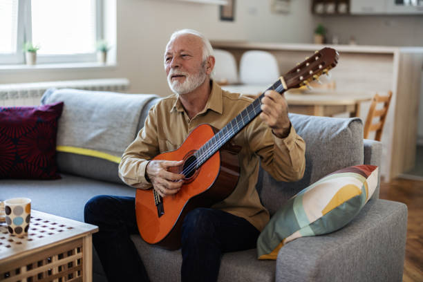 istock Bearded senior man with acoustic instrumental guitar. Man playing guitar while sitting on sofa in light living room. 2022320754