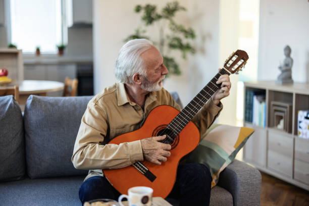 istock Bearded senior man with acoustic instrumental guitar. Man playing guitar while sitting on sofa in light living room. 2022322941