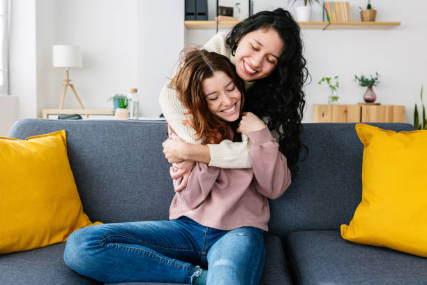 Two multiracial young women laughing and having fun together at home Two multiracial young women laughing and having fun together at home. Smiling latin american female hugging from behind her friendly girlfriend while sitting on sofa. Friendship concept lesbian couple embracing stock pictures, royalty-free photos & images