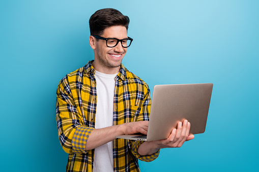 Photo of intelligent man with bristle dressed plaid yellow shirt in eyewear typing email on laptop isolated on blue color background.