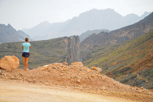 O beautiful woman in short skirt, tourist, taking photo  from the top of spectacular dirt road near Wadi Bani Awf—one of Oman’s most picturesque valleys.