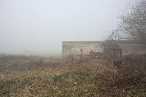 Boundary wall next to a creek with a weir in the italian countryside on a foggy day
