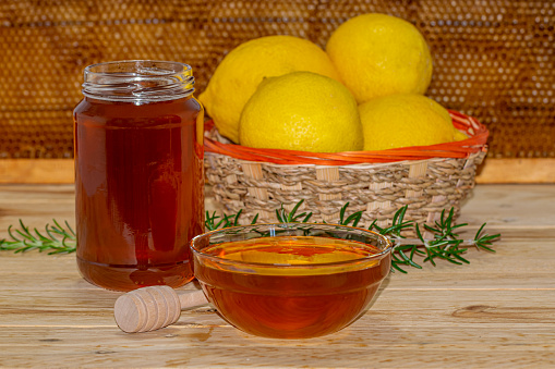 Honey jar without lid, dispenser, a bowl of honey, a rosemary spring and a basket of lemons on wood with a real honeycomb in the background