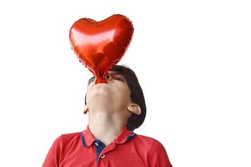 Horizontal head shot of cutout of one cute boy blowing or inflating fun love, valentine, anniversary theme red heart shaped foil balloon isolated over white transparent background, his head leaning or bending backwards with copy space