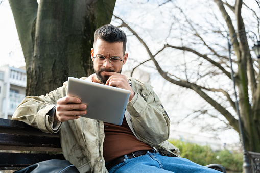 Young hipster millennial man world traveler nomadic living lifestyle sitting outdoor using digital tablet to search for new travel destination and places to visit. Generation Z expatriate male concept