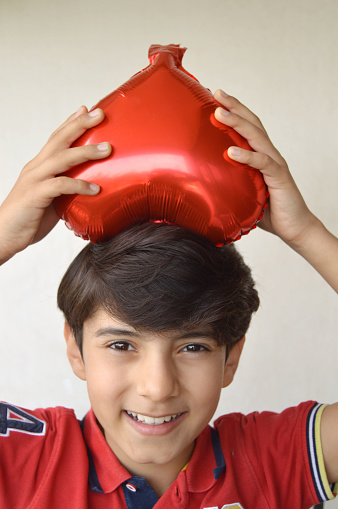 Vertical head shot of of one lovely happy smiling boy carrying one inflated fun love, valentine, anniversary theme red heart shaped foil balloon on head over grey background