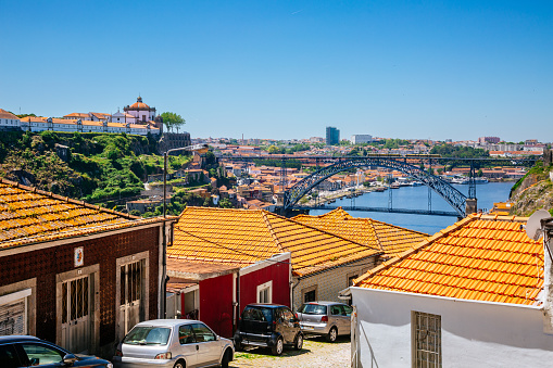 A sunny day in Porto, Portugal. In the foreground, you’ll find orange-tiled rooftops stretching across the frame. Along a narrow street, parked cars add a sense of scale. The iconic Dom Luís I Bridge stands out prominently, spanning the Douro River. Various buildings populate both sides of the river, and a hilltop church is visible. The clear blue sky is on the background.