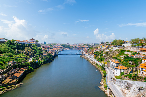A panoramic view captures the iconic Dom Luis I Bridge in Porto under a clear blue sky. The bridge spans the calm waters of the River Douro, connecting historic buildings and green landscapes on either side.