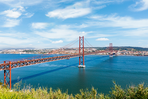 A panoramic view of a red suspension bridge - 25 de Abril Bridge extending over calm blue waters of Tagus river in Lisboa. In the foreground, lush green vegetation adds contrast. The cityscape forms the backdrop, with various buildings visible. Above the scene, a clear sky with a few clouds.