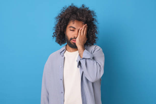 istock Young tired casual Arabian man millennial covers face with hand stands in studio 2022497570