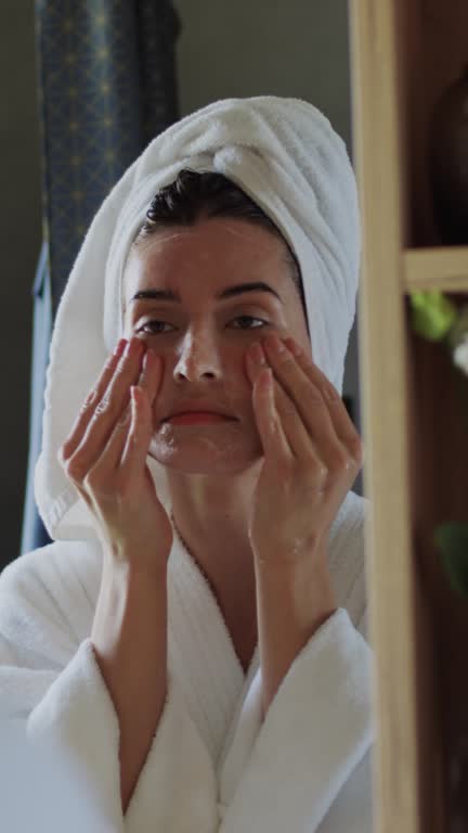 Young Woman with Hair in Towel Applying Skincare Treatment in front of Mirror