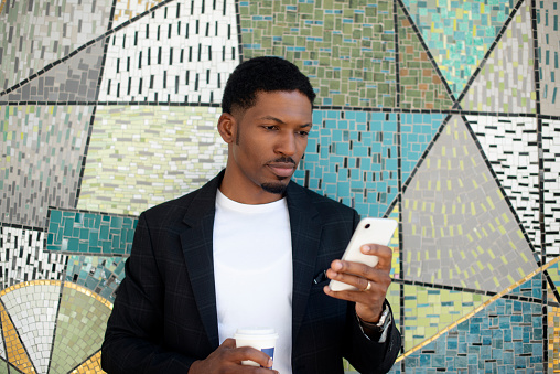 Phone, city and business black man on wall background for social media, networking and communication. Coffee, urban town and African person on smartphone for online chat on commute or travel in Kenya