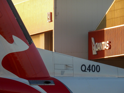 The vertical stabiliser and fuselage of a QantasLink De Havilland Canada Dash 8-400 plane, registration VH-QOA, parked at the Qantas Jetbase at Sydney Kingsford-Smith Airport.  In the background is an aircraft hangar. This image faces west and was taken from Qantas Drive, Mascot on a hot and sunny evening shortly before sunset on 17 February 2024.