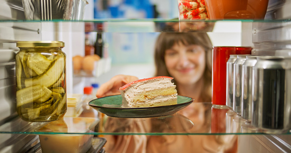 Smiling mature woman taking slice of cake out of refrigerator at kitchen.