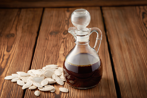 Glass decanter with pumpkin oil and a bunch of pumpkin seeds on a wooden background.