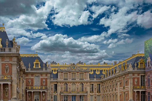 A stunning capture of the opulent exterior of Versailles Palace, adorned with golden ornaments and classical statues, set against a backdrop of a dramatic and expressive cloudy sky. The grandeur of the architecture is accentuated by the contrast between the intricate detailing of the building and the tumultuous weather above, showcasing the timeless elegance of this historic French landmark.