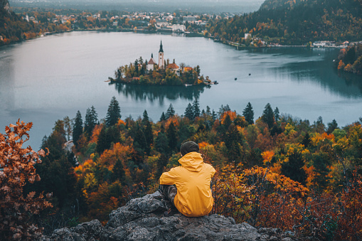 Rear view of a young traveler and adventurer dressed in a bright yellow raincoat, who finds solace and wonder amidst the famous alpine Bled Lago (Blejsko jezero) in Slovenia, on a foggy and rainy autumn day and finds serenity while sitting on a rock. From the vantage point of Mala Osojnica, the traveler gazes out at the mystical landscape, where the island with its picturesque church, the iconic Bled Castle, and the majestic Julian Alps are veiled in the ethereal embrace of fog and rain. This captivating scene encapsulates the spirit of exploration and appreciation for nature's ever-changing beauty, inviting viewers to embark on their journey of discovery in Slovenia's alpine paradise.