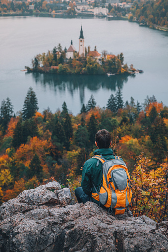 Rear view of a young traveler and adventurer man dressed in a raincoat and backpack looking at the view of the famous alpine Bled Lago (Blejsko jezero) with colorful trees in Slovenia, on a foggy and rainy autumn day and finds serenity while sitting on a rock. From the vantage point of Mala Osojnica, the traveler gazes out at the mystical landscape, where the island with its picturesque church, the iconic Bled Castle, and the majestic Julian Alps are veiled in the ethereal embrace of fog and rain.