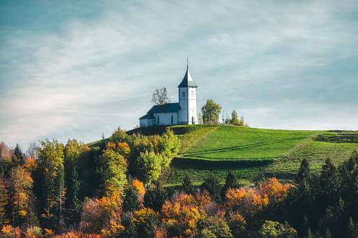 antastic charming Saint Primoz church, located atop a small hill in the picturesque Jamnik village of Slovenia, Europe. The church is beautifully lit by the golden hues of a stunning sunset, with the colorful mountain backdrop adding to the breathtaking scene. The photo was taken during the autumn season, and the trees surrounding the church are in full display of their warm and vibrant colorful fall colors.