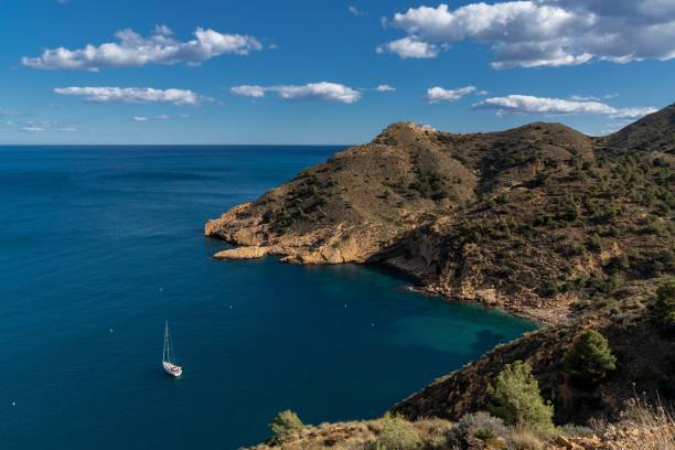 barca a vela ormeggiata in una baia del parco naturale della serra gelada con il faro di albir sullo sfondo - benidorm alicante spain headland foto e immagini stock