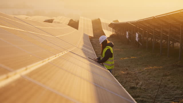 SLO MO Female Engineer in Protective Clothing Placing Digital Tablet on Solar Panel at Solar Power Station During Golden Hour