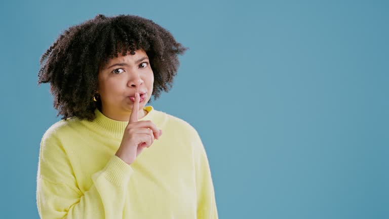 Woman, face and finger with quiet in studio for secret information or announcement, gossip or surprise. Female person, hand gesture and whisper for confidential news, blue background or mockup space