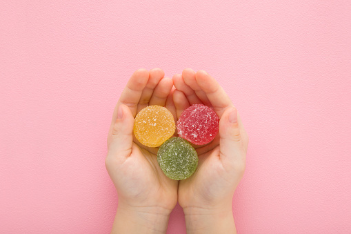 Sweet colorful candied fruit jelly with sugar grains in little child opened palms on light pink table background. Pastel color. Closeup. Point of view shot. Children sweets. Top down view.