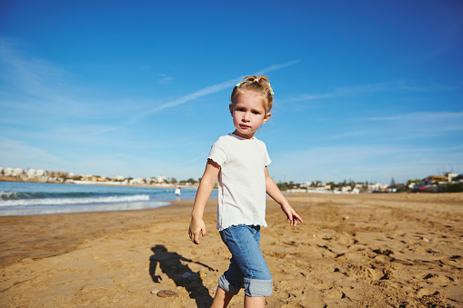 Adorable Caucasian blonde child girl in white t shirt and blue jeans, looking at the camera while walking barefoot on the wet sand, enjoying happy weekend with her family outdoor.