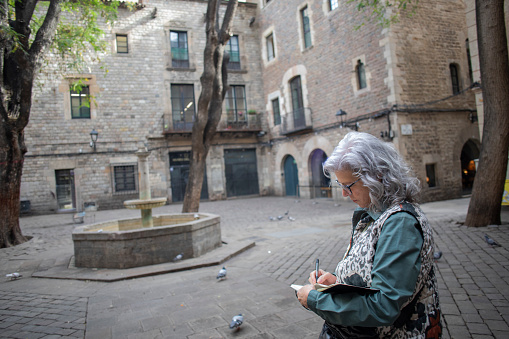 Lviv, Ukraine - 26 May 2019: Elderly woman in downtown. Old woman sitting on street, selling flowers and reading newspaper near house