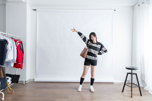 A young Asian woman standing in a studio with a clothes rack and stool, pointing to something off-camera