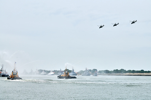 Fireboat sailing along Russian naval forces parade warships with military helicopters along coastline, seafaring tradition of military ships formation, nautical spectacle of russian sea power