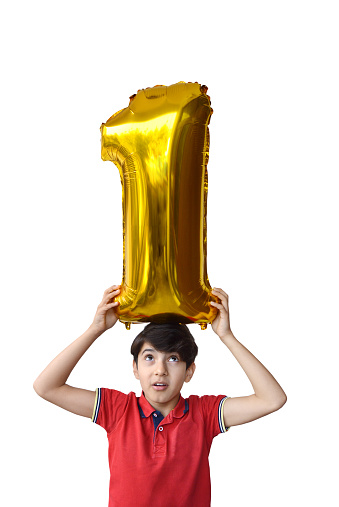 Cutout of waist up 11 year old tensed burdened child in red t shirt holding, carrying load of golden shiny 3 D number1 or one foil balloon on his head for position, success, win, 1st happy birthday celebrations isolated over vertical white background