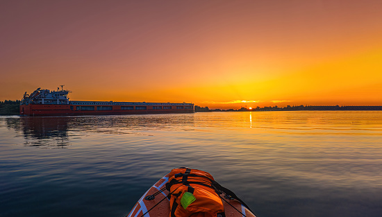 Hanging lifeboat, orange safety beacon and life preserver ring on the railing on the wooden promenade deck of a luxury cruise ship at dusk.  Small aperture creates star effect on the sun as it is peaking from behind the life preserver.  Boat is on a calm blue sea with no land in sight.