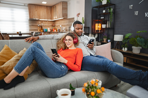 Happy multiracial couple with wireless headphones relaxing at home and using mobile phones