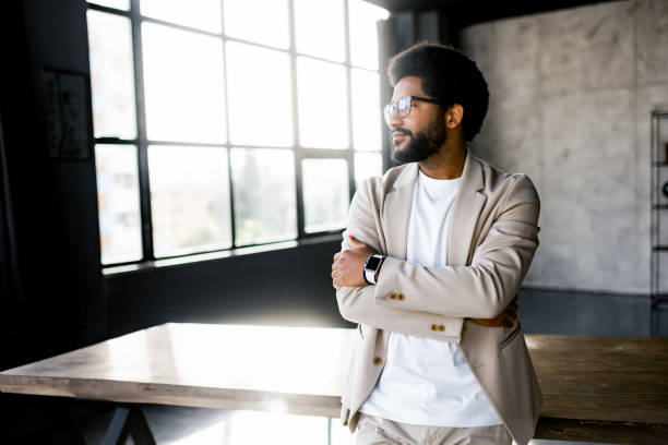 A cheerful Brazilian businessman in a smart casual beige suit in loft office A confident young businessman stands with arms crossed in a sunlit modern office, gazing thoughtfully out of large windows, embodying success and professionalism mid adult men stock pictures, royalty-free photos & images