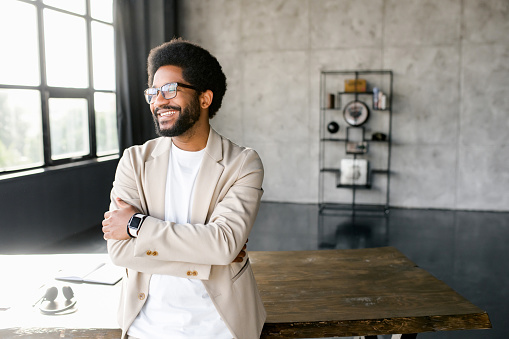 Young businessman in a beige suit strikes a pose with his arms confidently crossed, reflecting a persona of determination and professionalism. Male office employee looking aside with smile